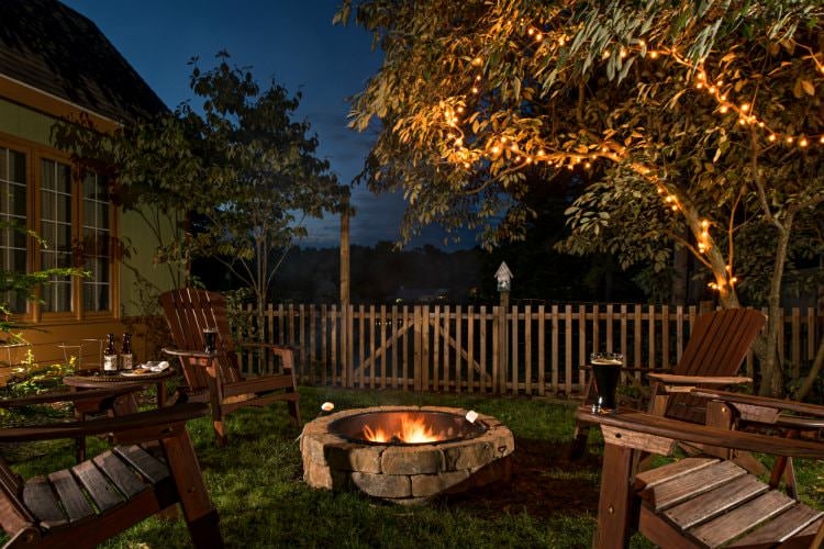 Cozy outdoor setting at nighttime, wooden lawn chairs surrounding a stone fireplace, twinkle lights in the trees