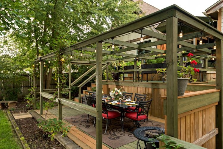 A wooden back porch dining area covered by a wooden Catalina surrounded by green foliage