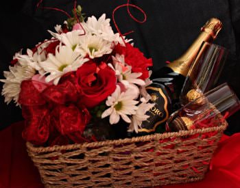 A tan wicker basket holding two wine glasses, a bottle of wine, a bouquet of flowers