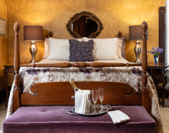 A bedroom with yellow antiqued walls, a four poster bed, a purple cushioned sitting bench at the foot of the bed with a silver tray with a bucket of champagne and 2 champagne flutes on it.