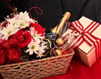 A tan wicker basket holding two wine glasses, a bottle of wine, a bouquet of flowers and a present