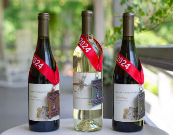 Three bottles of wine sitting on whit table with red ribbon showing 2024 holding a medallion