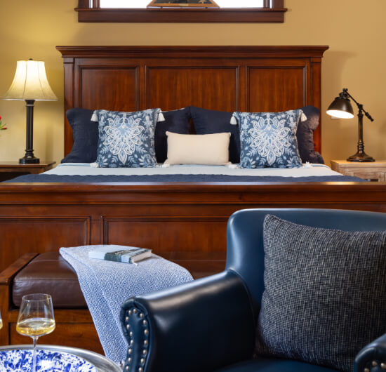 A bedroom with a bed with tall wood headboard and footboard, blue and white bedding, a leather sitting bench at the foot of the bed, and a blue leather armchair and a small table with a blue plate and glass of white wine.