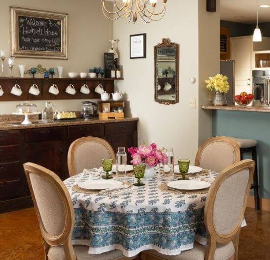 A dining room with a table and tablecloth, plates, green glasses, 4 chairs, a buffet table with food and coffee cups, and a breakfast bar with barstools
