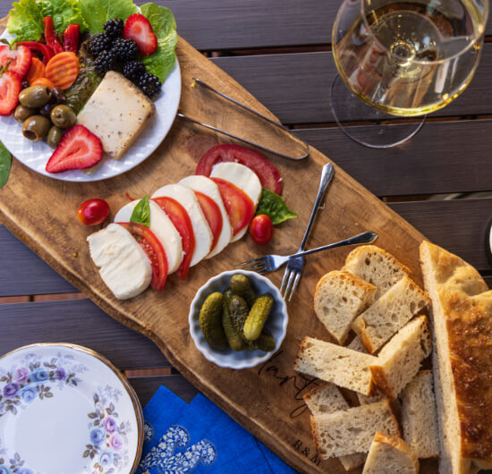 A wood charcuterie board with cheese, fresh fruits and vegetables, pickes, olives, and breads, along with some chine plates and a glass of white wine on either side.