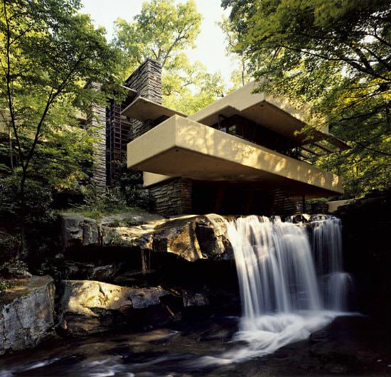 A modernized grey stone building built at the edge of a waterfall and stream, surrounded by thick foliage and bushes