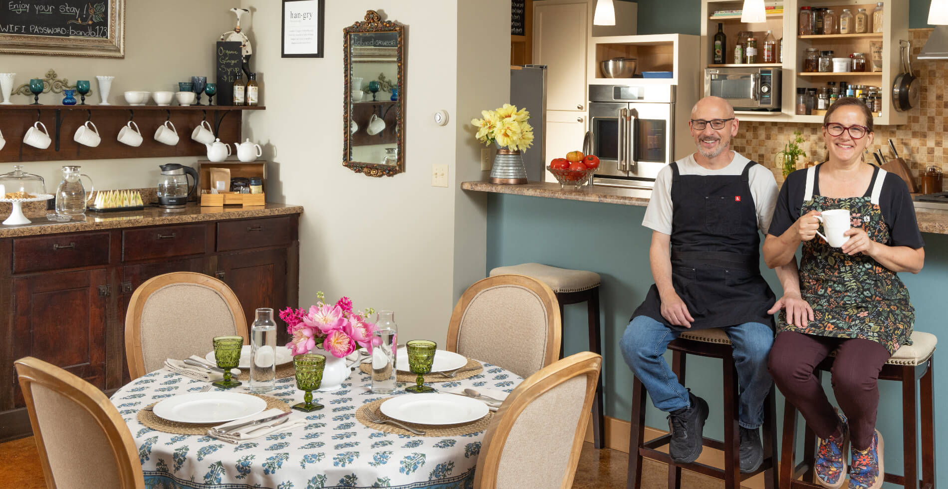 A dining room with a table set for a meal, a breakfast bar along one wall, a smiling couple wearing aprons seated at some bar stools in front of a breakfast bar with a kitchen behind them.