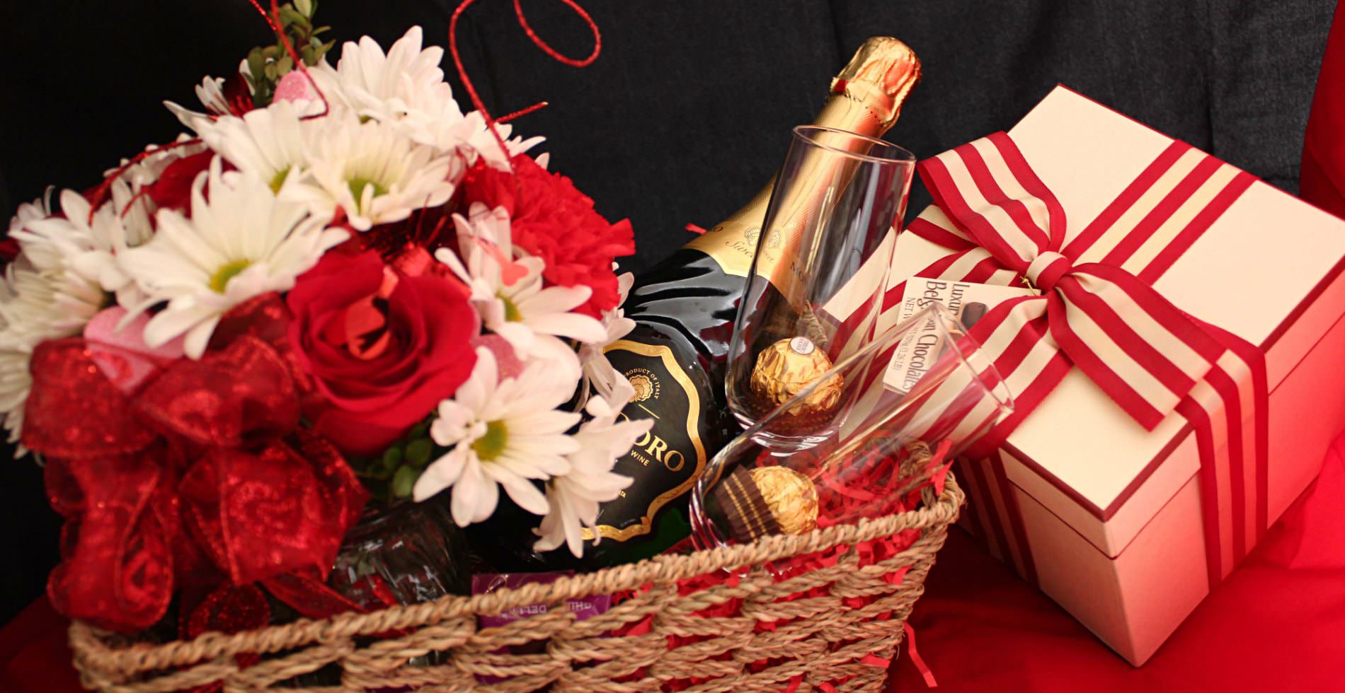 A tan wicker basket holding two wine glasses, a bottle of wine, a bouquet of flowers and a present