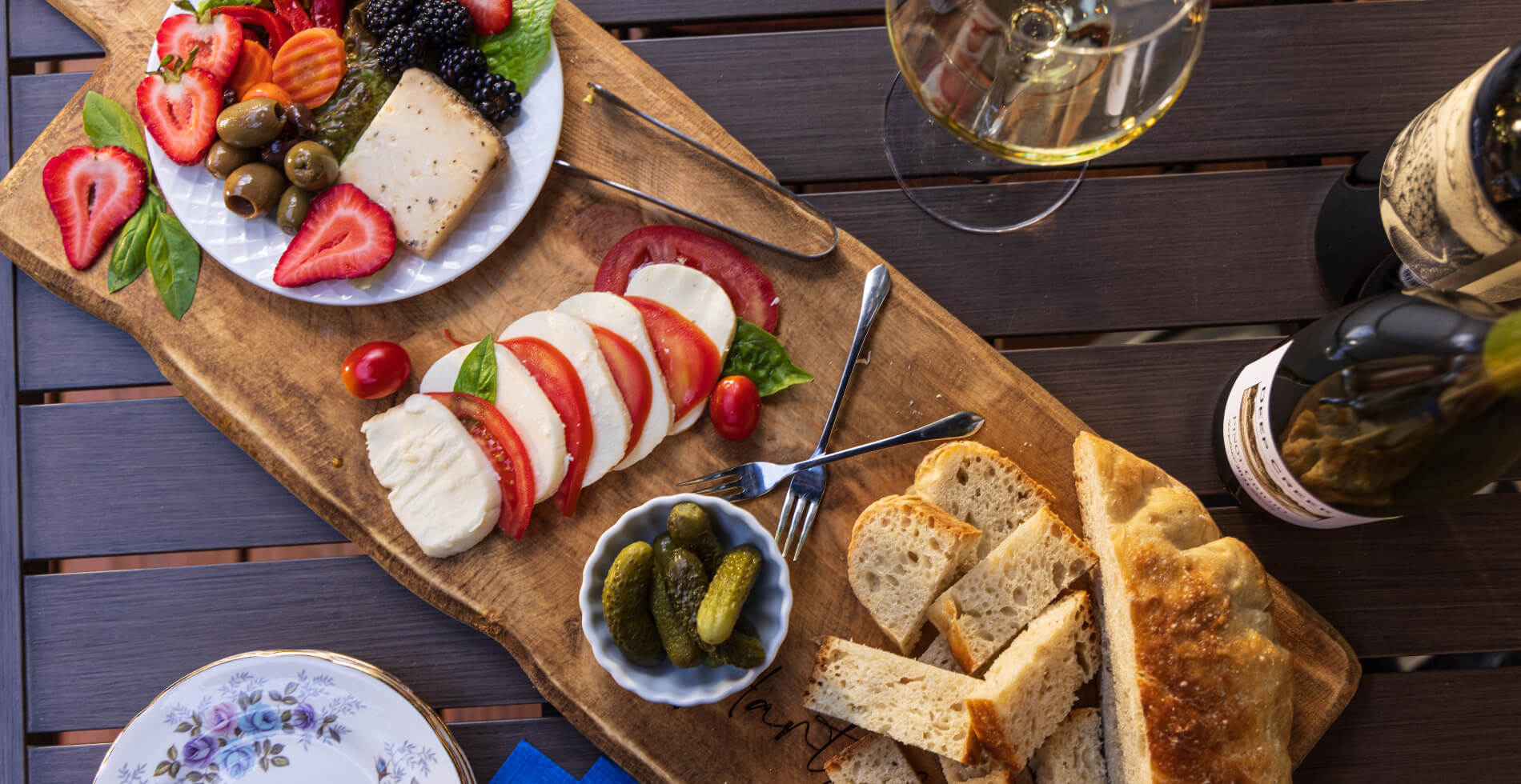 A wood charcuterie board with fresh fruits and vegetables, cheeses, bread, along with a bottle of wine with a wine glass with white wine, and china plates on the side.