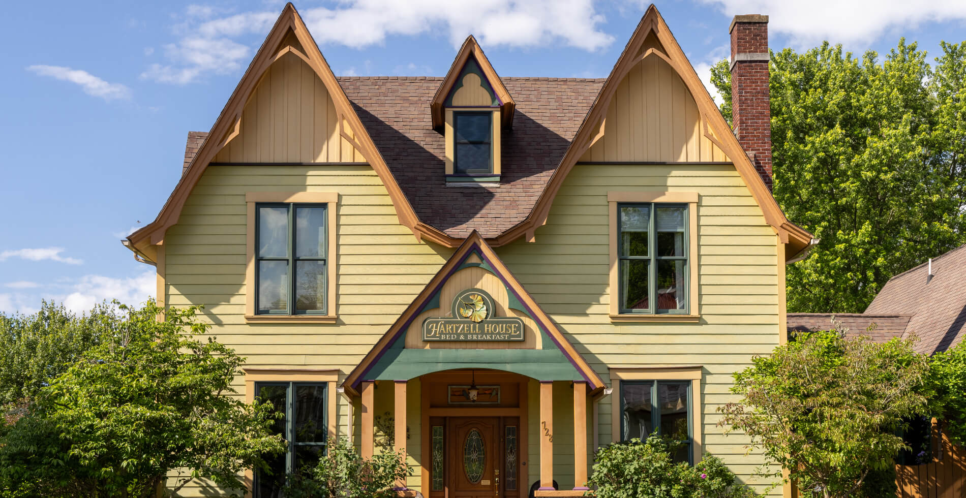 A charming yellow Victorian home with dormers and a sign above the front door that says Hartzell House Bed & Breakfast, surrounded by bushes and trees.