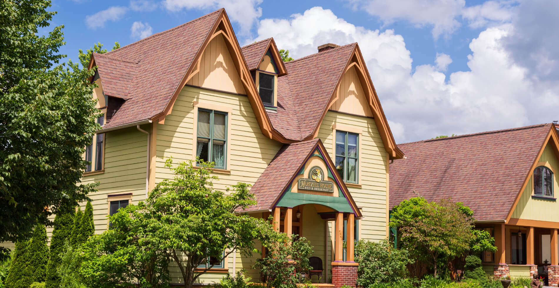A yellow house with dormers and pitched roofs with Hartzell House Bed & Breakfast above the front door, surrounded by lush trees and bushes with another similar house next to it.