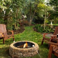 Outside view of a grassy backyard, several lawn chairs around a stone fireplace, a green path leading away from scene