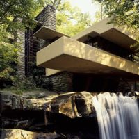 A modernized grey stone building built at the edge of a waterfall and stream, surrounded by thick foliage and bushes