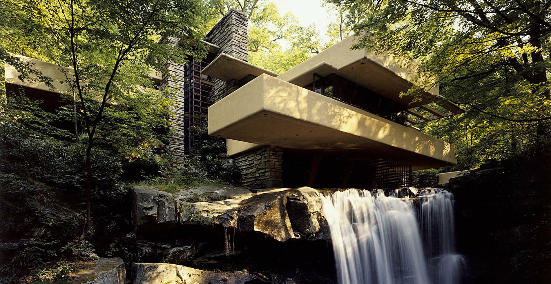 A modernized grey stone building built at the edge of a waterfall and stream, surrounded by thick foliage and bushes