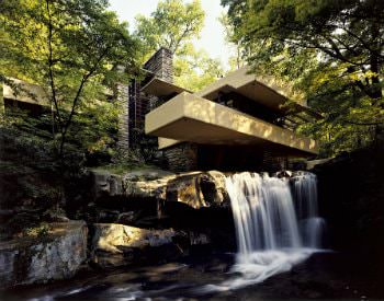 A modernized grey stone building built at the edge of a waterfall and stream, surrounded by thick foliage and bushes