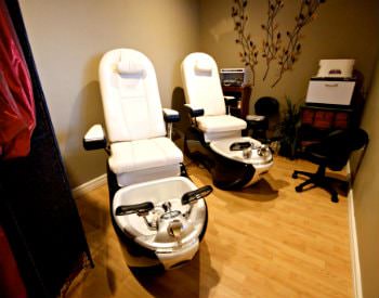 An interior view of a massage room, two white chairs with feet soaking tubs and taupe walls