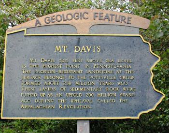 A slate colored sign that says, “A geologic feature, MT. Davis”, mounted on a wooden post surrounded by green shrubbery.