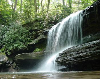 A gorgeous waterfall cascading over smooth and jagged rocks into a bed of water, surrounded by looming green trees