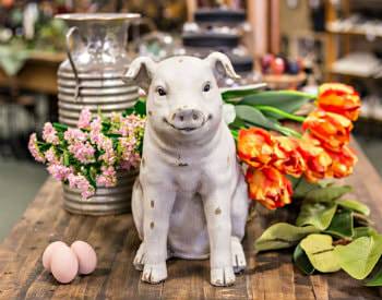 A stone pig sculpture sitting on a brown wooden table surrounded by flower bouquets and 3 eggs