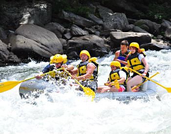 A grey river raft going down strong river rapids with a group of people wearing yellow safety garb