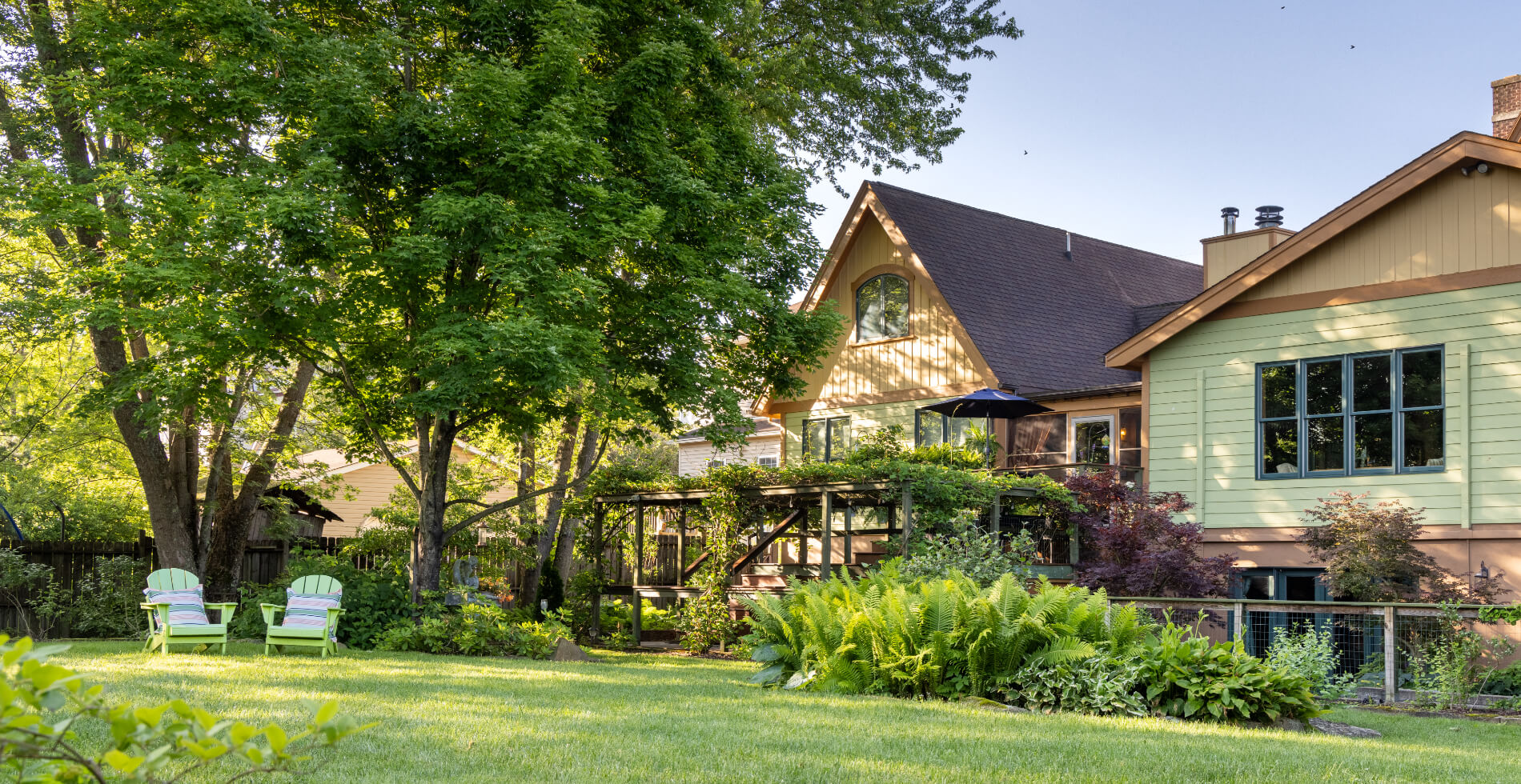 A large grassy yard with 2 green adirondack chairs under mature shade trees, lush bushes and and a sprawling yellow house.